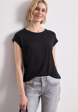 Femme blonde souriante, portant un t-shirt noir à manches courtes et un jean gris clair, debout contre un mur blanc uni.