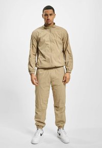 Beige lettvekts treningsdrakt med glidelåsjakke og elastiske mansjetter. Glatt tekstur, tettsittende bukser, brukt med hvite joggesko.