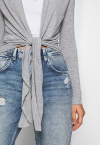 Grå omslagströja knuten i midjan, parat med slitna blå jeans med ljusa tvättar och trasiga fållar.