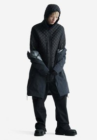 Giacca trapuntata nera con cappuccio, indossata sopra un lungo cappotto nero. Abbinata a pantaloni neri e stivali neri robusti. Texture liscia.