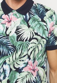 Koszulka polo z krótkim rękawem z granatowym kołnierzykiem, ozdobiona wzorem tropikalnych liści i kwiatów hibiskusa w odcieniach zieleni i różu na ciemnym tle.