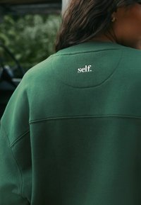 Groene sweatshirt met een ronde pasvorm en een gestikte detail, met het woord "self." in het wit geborduurd op de achterkant. Zachte textuur.