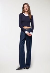 Top lungo a maniche corte di colore blu navy con scollatura a V indossato sopra una camicia bianca con colletto, abbinato a jeans in denim scuro a gamba larga e tacchi neri a punta con decorazioni.