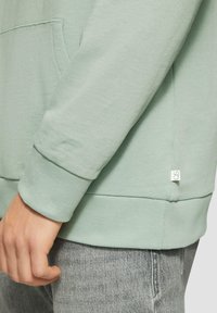 Felpa verde chiaro con tasca a marsupio, polsini e orlo a costine. Presenta un piccolo etichetta sul lato con un semplice logo. Il tessuto è morbido e texturizzato.