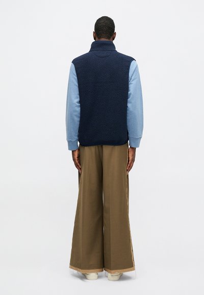 Námornícky fleecový vest s vysokým golierom, vrstvený nad svetlomodrou košeľou s dlhým rukávom. Voľné hnedé nohavice so širokými nohavicami a biele tenisky.