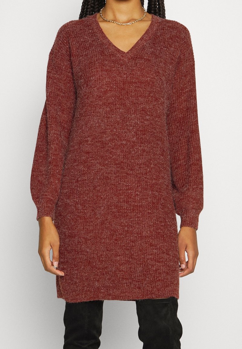Femme portant une robe pull en maille rouge brique à manches longues avec un col en V et un pantalon noir, debout devant un fond clair uni.