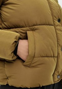 Kurtka puchowa w kolorze oliwkowym, z pikowanym wzorem, z zapięciem na guziki i kieszeniami bocznymi. Miękki materiał zapewnia izolację i ciepło.