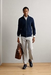 Mornarska zip-up jopica, bela srajca s ovratnikom, svetlo sivi hlače, črne superge, drži rjavo usnjeno torbo. Lesena tla, nevtralen ozadje.