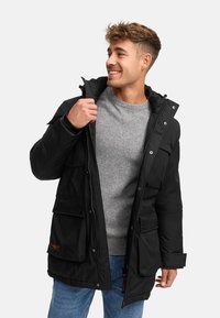 Giacca nera imbottita con chiusura a zip e bottoni automatici, dotata di più tasche frontali e cappuccio con cordino, indossata sopra un maglione grigio.