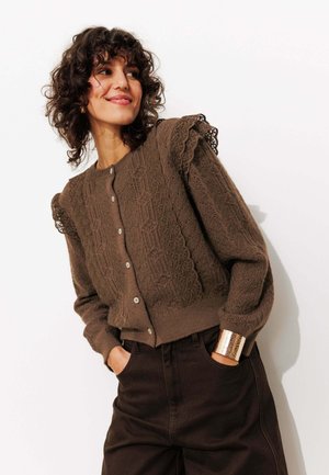 Cardigan en tricot marron avec des motifs texturés, des accents en dentelle sur les manches et une fermeture à boutons. Assorti à un pantalon sombre et un bracelet métallique.