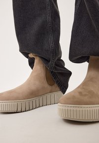 Bottines en suede beige avec des panneaux élastiques sur les côtés et des semelles en caoutchouc texturé, assorties à un pantalon en denim foncé ample.