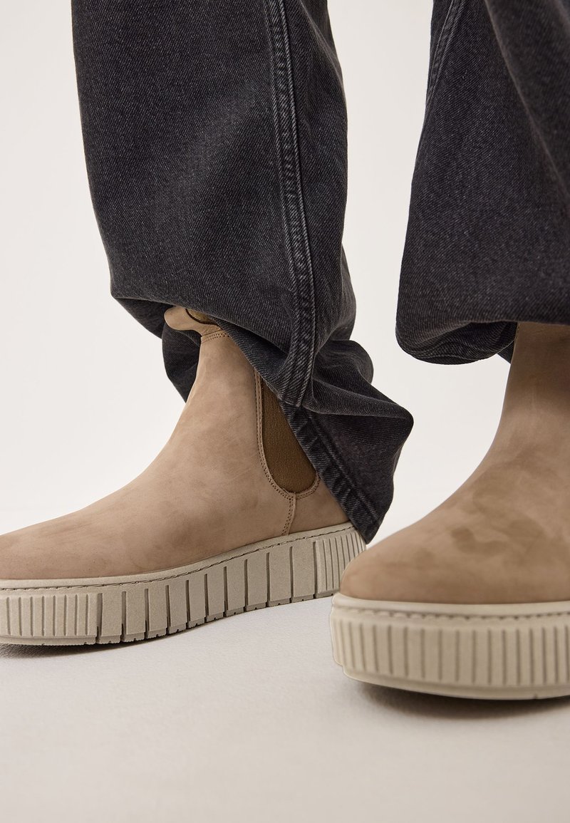 Bottines en suede beige avec des panneaux élastiques sur les côtés et des semelles en caoutchouc texturé, assorties à un pantalon en denim foncé ample.