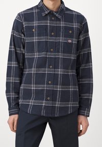 Camicia di flanella a quadri blu navy con strisce bianche, due tasche sul petto, chiusura con bottoni e bottoni marroni. Texture morbida e design del colletto classico.