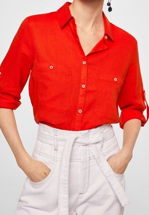 Nainen, joka pukeutunut kirkkaan punaiseen napitettuun paitaan, jossa hihat käärittynä, sekä korkeavyötäröisiin valkoisiin housuihin, joissa on kangasvyö, kädet taskuissa.