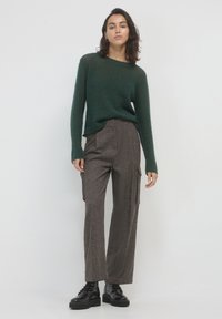 Frau in einem dunkelgrünen Strickpullover, braunen weiten Hosen mit Seitentaschen und schwarzen Schnürstiefeln, die vor einem schlichten weißen Hintergrund steht.