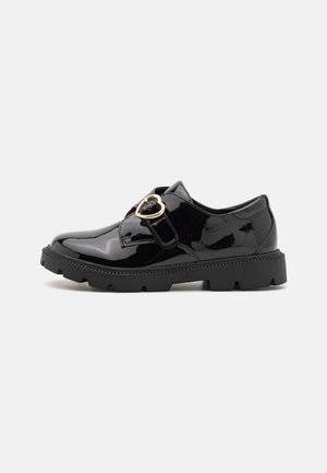 Los zapatos de charol negro presentan una puntera redondeada, un cierre lateral con un acento de corazón, una suela de goma texturizada y un tacón plano.