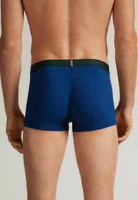 Modré trenýrky s zeleným elastickým pasem, s hladkou strukturou a přiléhavým střihem, viděné zezadu.