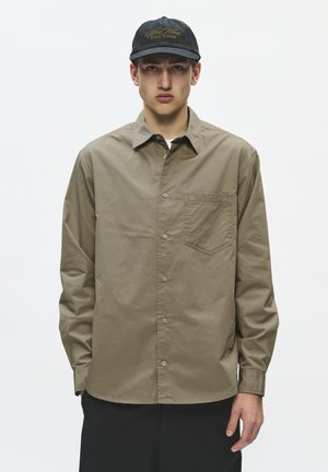 Jeune homme portant une chemise kaki boutonnée avec une poche poitrine et une casquette noire avec un texte jaune, debout devant un fond blanc uni.