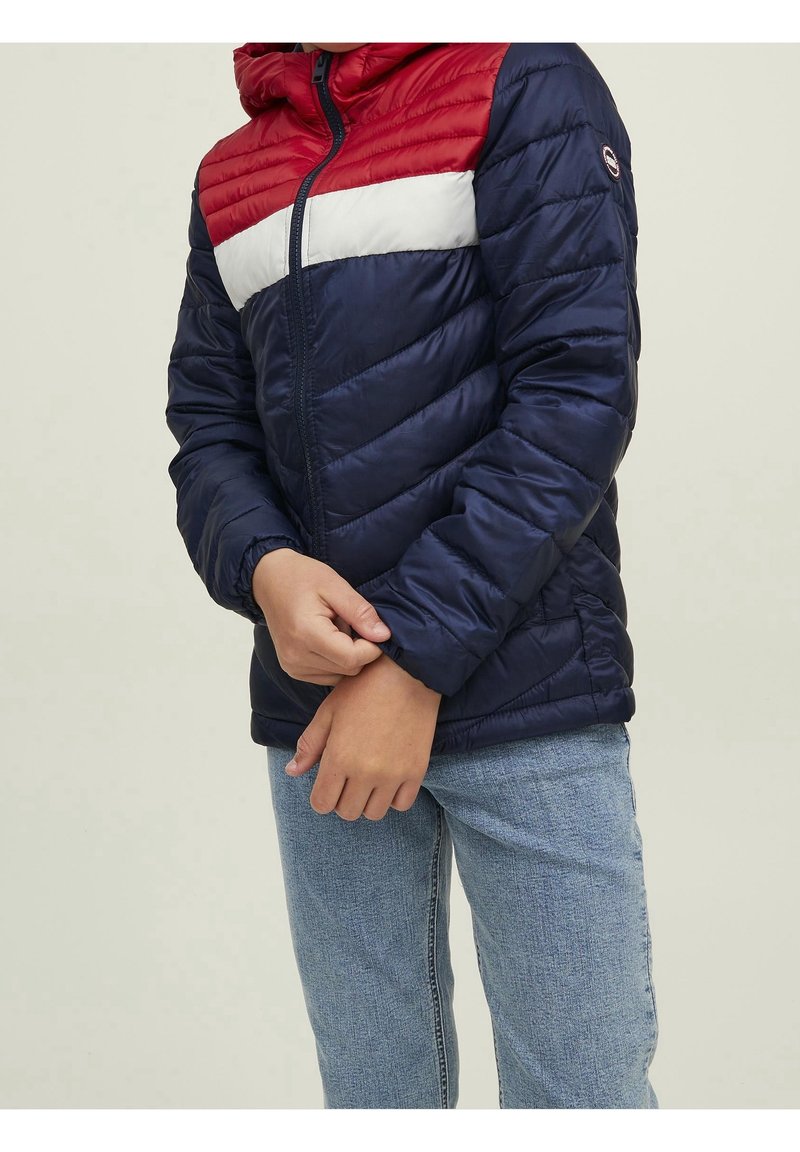 Vadderad jacka i marinblått med röda och vita horisontella ränder över bröstet. Har en huva och elastiska ärmslut. Materialet är slätt och glänsande.