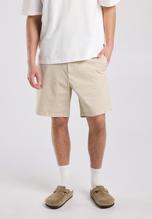 Mann trägt beige Shorts, weiße Socken, braune Slipper und ein lockeres weißes Kurzarmhemd, steht mit einer Hand in der Tasche.