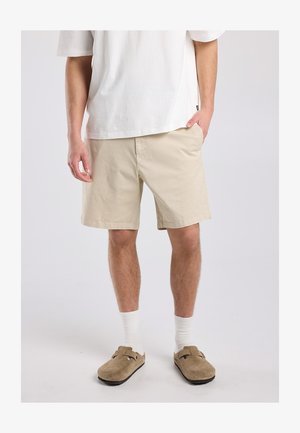 Mann trägt beige Shorts, weiße Socken, braune Slipper und ein lockeres weißes Kurzarmhemd, steht mit einer Hand in der Tasche.