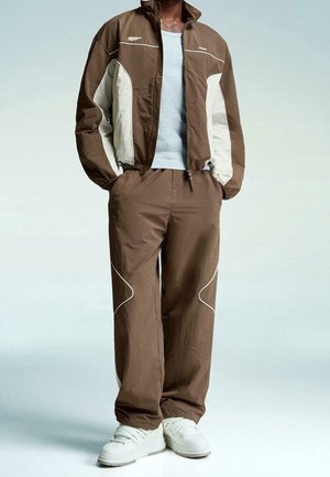 Person iført brun og beige vindjakke med matchende bukser og hvide sneakers, stående med hænderne i lommerne.