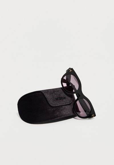 Γυαλιά ηλίου με μαύρους φακούς σε σχήμα γάτας, τοποθετημένα πάνω σε μαύρη θήκη με υφή, πάνω σε ανοιχτό φόντο.