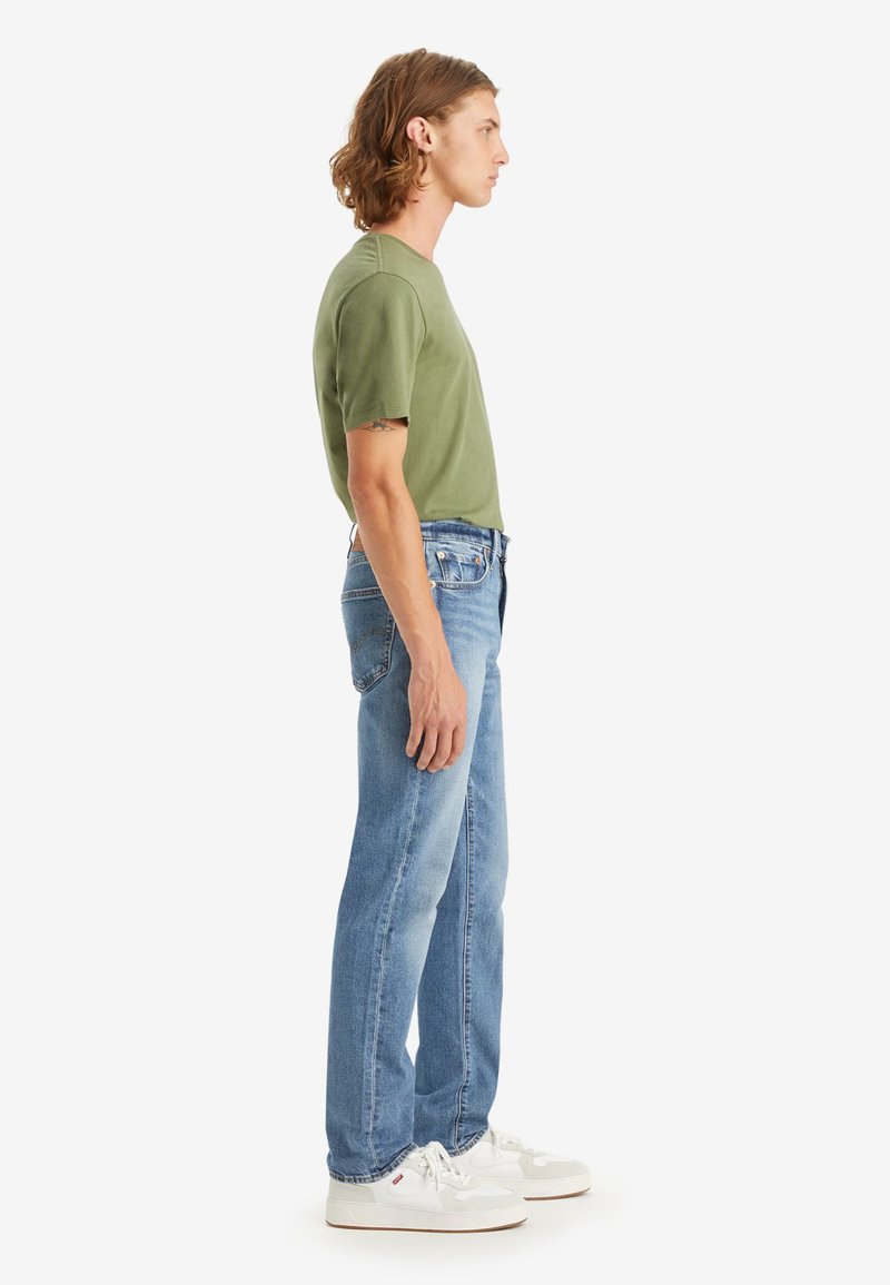 Camiseta verde oliva, de mangas cortas, combinada con vaqueros de mezclilla azul claro. Las zapatillas son blancas con acentos beige. Vista de perfil lateral.