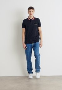 Μαύρο polo μπλουζάκι με κόκκινο και λευκό ριγωτό γιακά, συνδυασμένο με μπλε τζιν παντελόνι και λευκά αθλητικά παπούτσια, που παρουσιάζεται σε ουδέτερο φόντο.