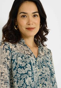 Blouse florale en teal et crème, avec un col, une fermeture à boutons et une texture transparente ornée de motifs complexes et de détails froncés.