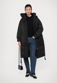 Long manteau en duvet noir avec un col haut, texture matelassée et ourlet à cordon de serrage. Assorti à un pull sombre et à un jean ample bleu.