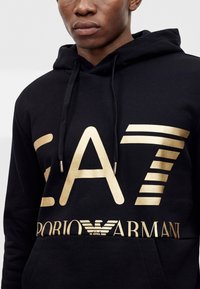 Sweat à capuche noir avec logo doré EA7 Emporio Armani porté par une personne au visage neutre, cadré à la bouche et à la poitrine.