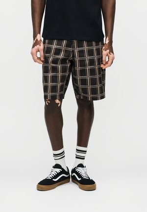 Persona con pantalones cortos de cuadros negros y beige, camiseta negra, calcetines blancos con rayas negras y zapatillas negras con cordones blancos, de pie sobre fondo blanco.