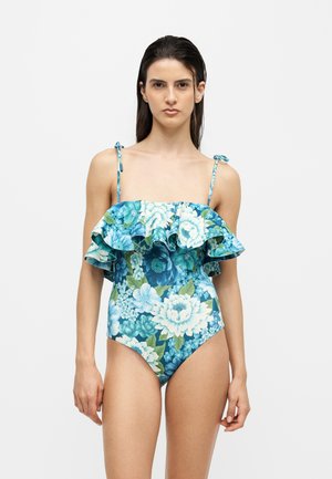 Γυναίκα με μπλε ολόσωμο μαγιό με floral σχέδιο, βολάν και λεπτές τιράντες, στέκεται μπροστά σε απλό φόντο.