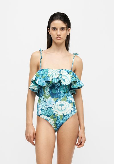 Femme portant un maillot de bain une pièce bleu à motif floral avec un design épaules dénudées à volants et des fines bretelles nouées, debout devant un fond uni.