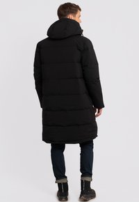 Chaqueta de plumas negra con capucha, que presenta una silueta larga, un diseño acolchado y una textura suave y mate. Combinada con jeans oscuros y botas.