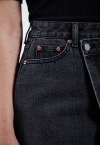 Zwarte denim jeans met een hoge taille, voorzien van koperen klinknagels, een zilveren knoop en een gestructureerde stof. Close-up van de zakken en de tailleband.