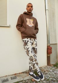 Jeune homme appuyé contre un mur beige, portant un sweat à capuche marron avec un motif graphique, un pantalon en polaire blanc avec des motifs de feuilles foncées et des baskets noires.