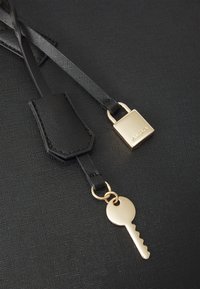 Schwarzer strukturierten Stoff mit einem goldenen Vorhängeschloss und einem Schlüsselakzent. Der Schlüssel hat eine klassische Form, während die Riemen glatt mit einem leichten Glanz versehen sind.