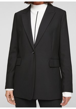 Blazer noir cintré à bouton unique porté sur un top blanc à col montant avec une bande noire verticale au centre, porté par une personne.