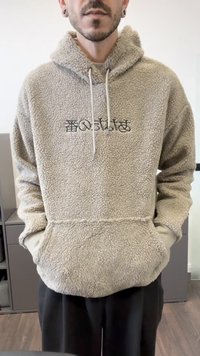 Man in een beige fleece hoodie met zwarte geborduurde tekst, met zijn handen in de voorzak, staand binnen op een houten vloer.