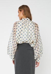 Blusa leggera di colore crema con pois blu, dotata di un colletto alto e maniche ampie e trasparenti. Abbinata a una gonna grigia.