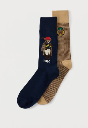 Chaussette marine avec un ourson en tenue polo accompagné du texte "POLO", associée à une chaussette beige arborant un motif à carreaux marron et un blason brodé.