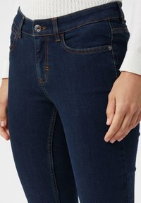 Donkerblauwe denim jeans met vijf zakken, oranje stiksels en een zilveren knoopsluiting. De stof heeft een gladde, licht gestructureerde afwerking.