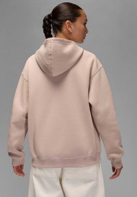 Przesadzony różowy hoodie z kapturem na sznurki, prążkowanymi mankietami i dołem. Gładka tekstura z minimalnymi detalami szycia. Noszony z białymi spodniami.