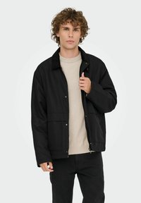Ung mand med krøllet hår iført en sort zip-up jakke over en lys beige sweater og sorte bukser, mens han holder om jakkens krave.
