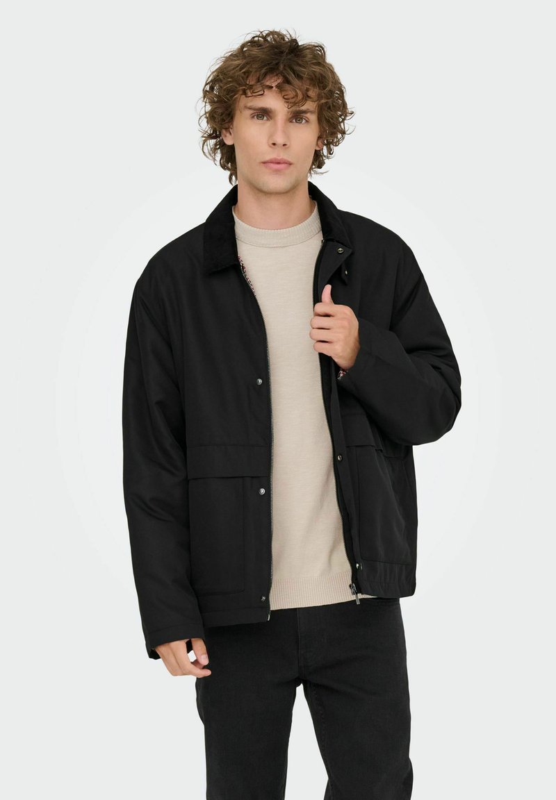 Ung mand med krøllet hår iført en sort zip-up jakke over en lys beige sweater og sorte bukser, mens han holder om jakkens krave.