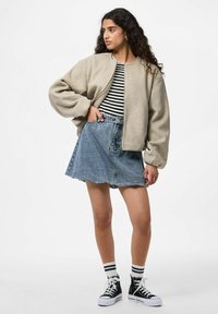 Vrouw met lange krullend haar, gekleed in een beige bomberjack over een zwart-witgestreept shirt, spijkerrok, gestreepte sokken en zwarte hoge sneakers.