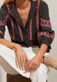 Blouse gris foncé avec broderie florale colorée, col à volants et poignets de manches. Pantalon blanc et bottines beige clair portés.