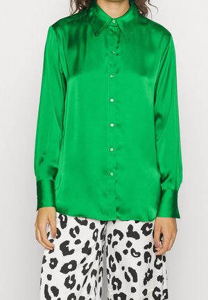 Persoon draagt een glanzend groen overhemd met lange mouwen en witte broek met zwart dierenprint, staand met armen ontspannen langs het lichaam.